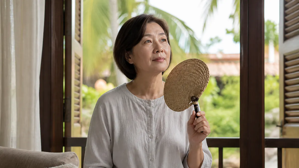 Malaysian Chinese woman cooling herself with a hand fan by a window.