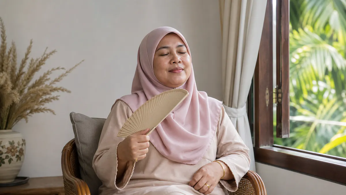 Wanita Melayu menyejukkan diri dengan kipas tangan di tepi tingkap.